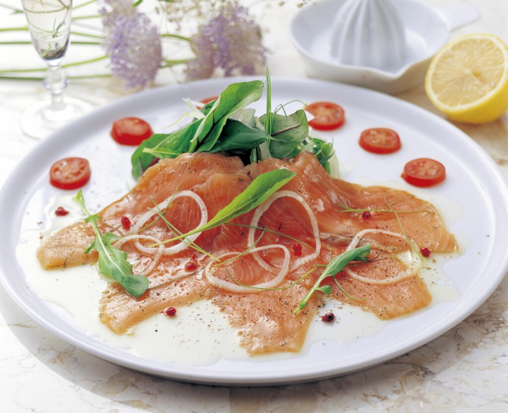 Carpaccio di salmone posizionato su di un piatto bianco e decorato con fettine di arancia sottili, rucola e olio condibio agli agrumi.