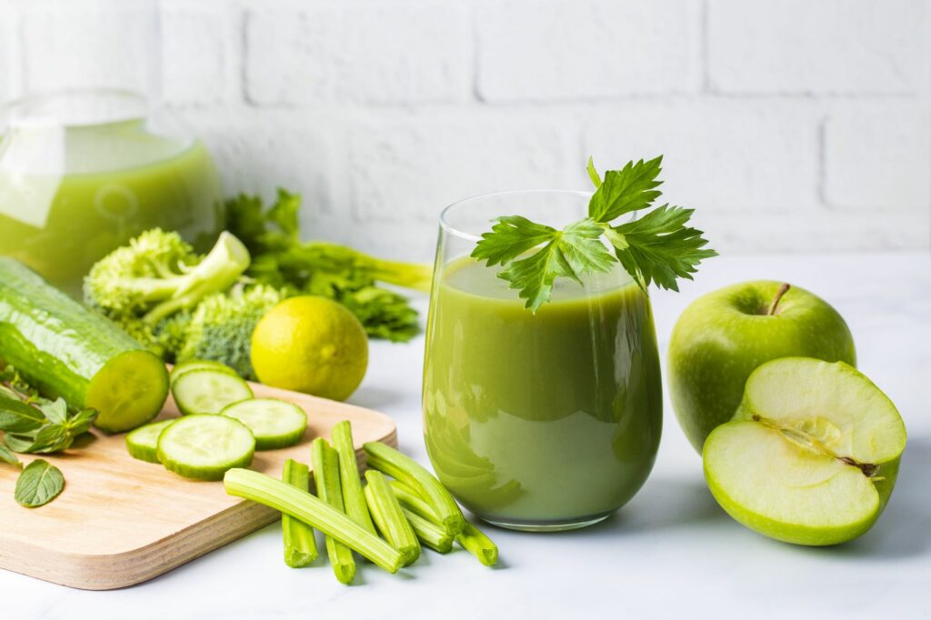 Smoothie verde in un bicchiere circondato da mele, cetrioli, e altre verdure verdi.